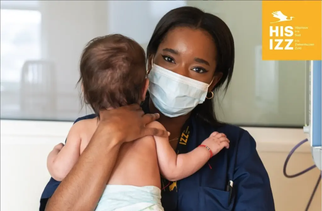 Healthcare professional wearing a face mask holding a baby in a hospital setting.