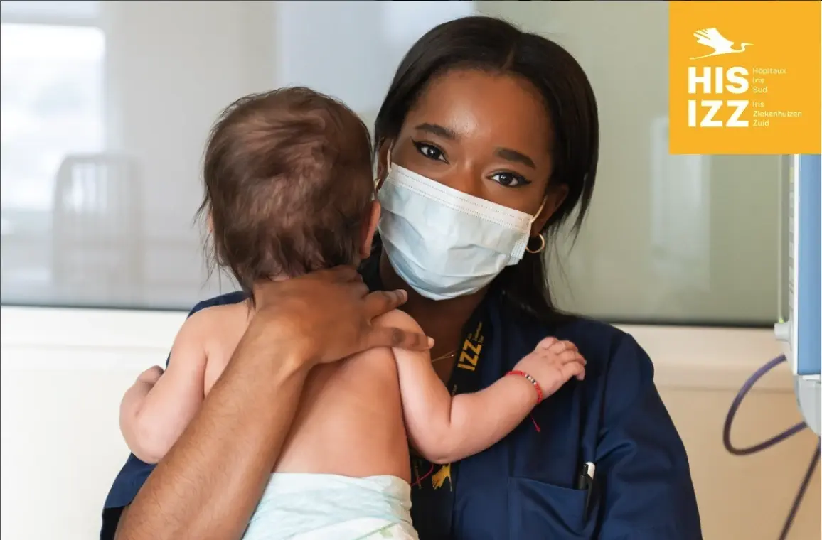 Healthcare professional wearing a face mask holding a baby in a hospital setting.